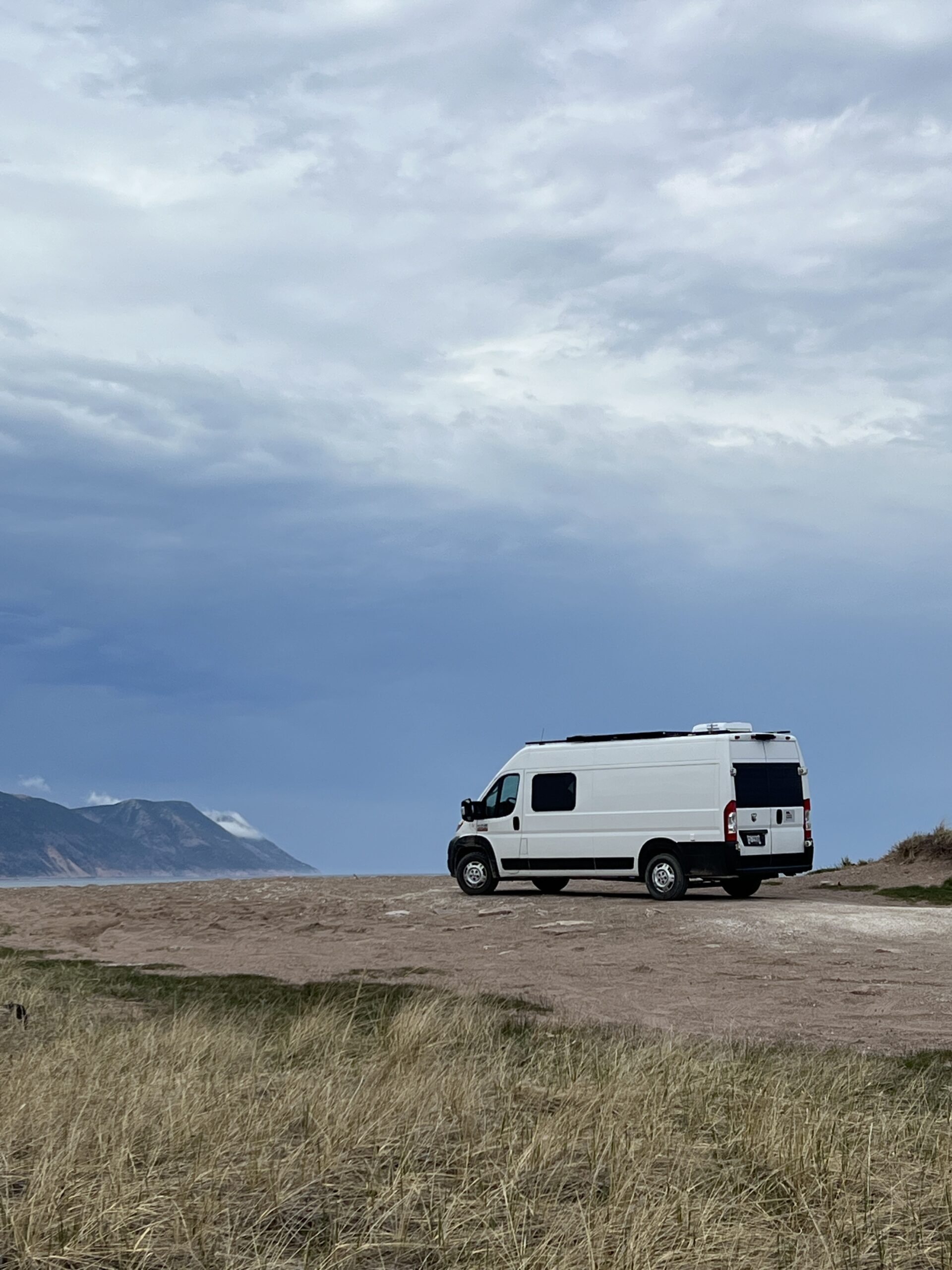 A white camper van parked on a beach, with mountains wrapped in cloud and the ocean in the background